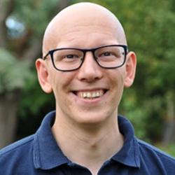 Bald man with glasses smiling into camera, head and shoulders shot. Trees out of focus in background