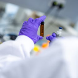 Purple gloved hand in white lab coat holding a brown glass vial