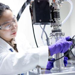 Woman with safety glasses, white lab coat and purple plastic gloves working with lab equipment/machine